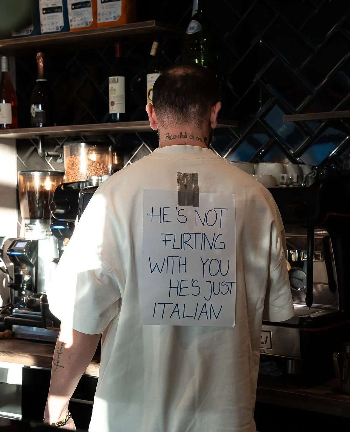 Pasquale von hinten an der Kaffeemaschine; auf seinem Rücken klebt ein Zettel: „He's not flirting with you, he's just Italian“.