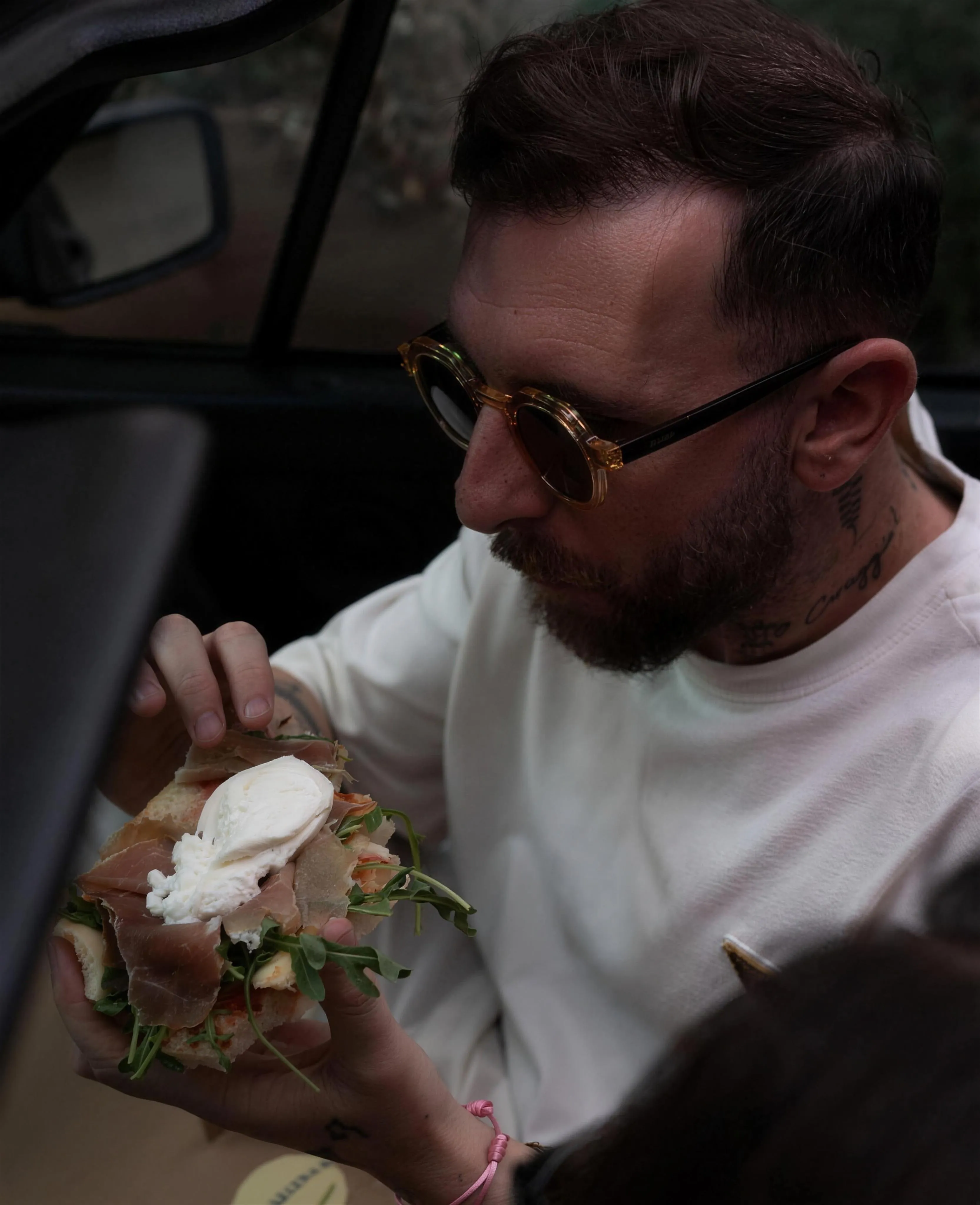 Pasquale sitzt im Auto, trägt eine runde Sonnenbrille und hält ein Stück Pizza.